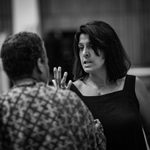 A black-and-white photo of a woman with shoulder-length dark hair and a black sleeveless top, in an animated conversation with a person whose back is facing the camera.