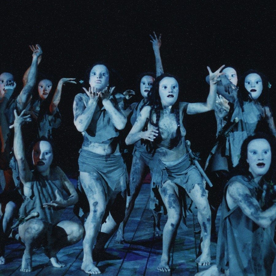 Performers on stage wear tattered, monochromatic costumes and eerie, white masks, their bodies contorted in expressive, dramatic poses. The lighting is dark, creating a haunting and theatrical atmosphere.