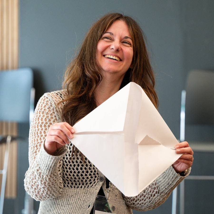 A woman with long brown hair, wearing a crocheted beige sweater, is smiling broadly while holding up an empty white piece of paper, folded like an envelope. She is standing indoors, with grey and beige furniture in the background, resembling a rehearsal room.