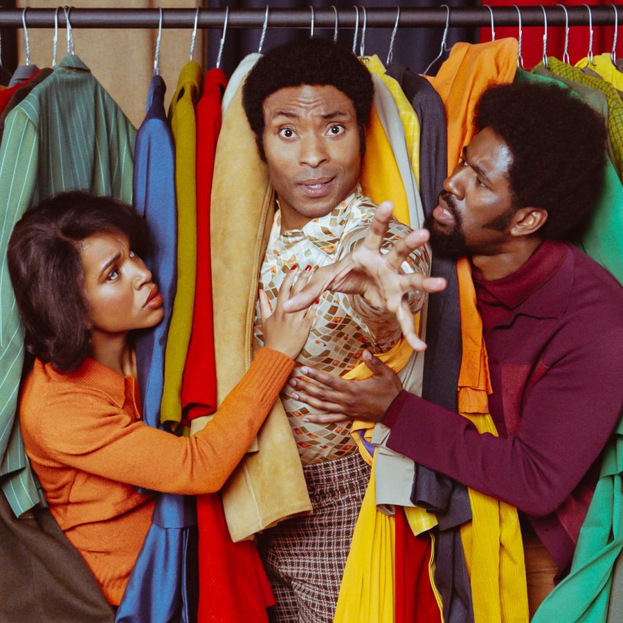 Three people are playfully interacting among colorful clothes hanging on a rack. The person in the center is being playfully grabbed by the others, creating a humorous scene against a backdrop of various vibrant garments.