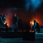 A dimly lit scene shows four people in heavy coats, standing and sitting amidst pottery.