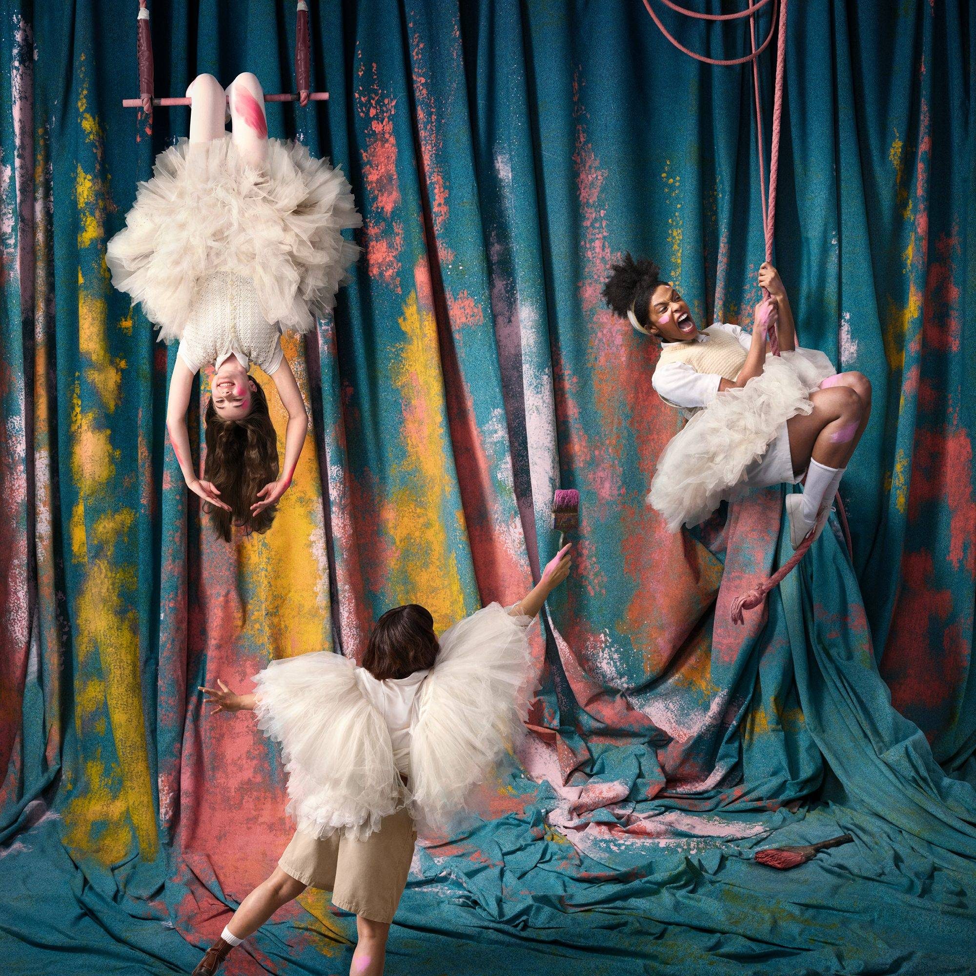 Three young women in white tulle dresses create dynamic poses against a colorful, textured curtain background. One hangs upside down, another swings on a rope, and the third stands below reaching up. The scene is vibrant and whimsical, evoking a sense of playful elegance.