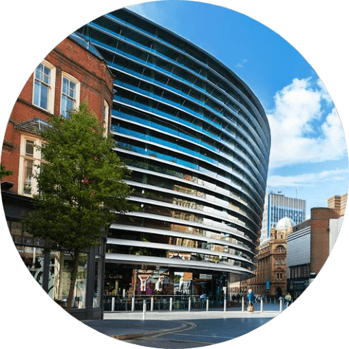 A modern, curved, glass-fronted building with horizontal slats stands prominently in an urban setting under a blue sky with scattered clouds. Surrounding the building are older, brick structures, trees, and a partially visible high-rise in the background.