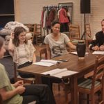 Four people are seated around a wooden table in a rehearsal room, engaged in conversation and laughing. The table has an ornate urn and rehearsal scripts. In the background is a clothes rail with coats, stacked furniture, and household items.