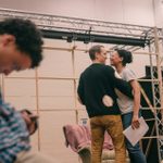 In the foreground, a person in a blue checkered shirt is sitting and looking at a phone. In the background, two people are standing and laughing together, one holding a rehearsal script. They are in a rehearsal room with minimal stage rigging behind them.