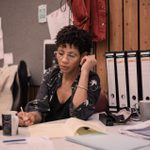 A person with short curly hair and glasses is seated at a cluttered desk, engrossed in writing on a rehearsal script. They are surrounded by various folders, papers, and a few mugs.