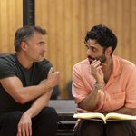 Two men are engaged in a deep conversation. The man on the left gestures with his hand while the man on the right holds a pen and an open book. They are sitting against a backdrop of wooden paneling.