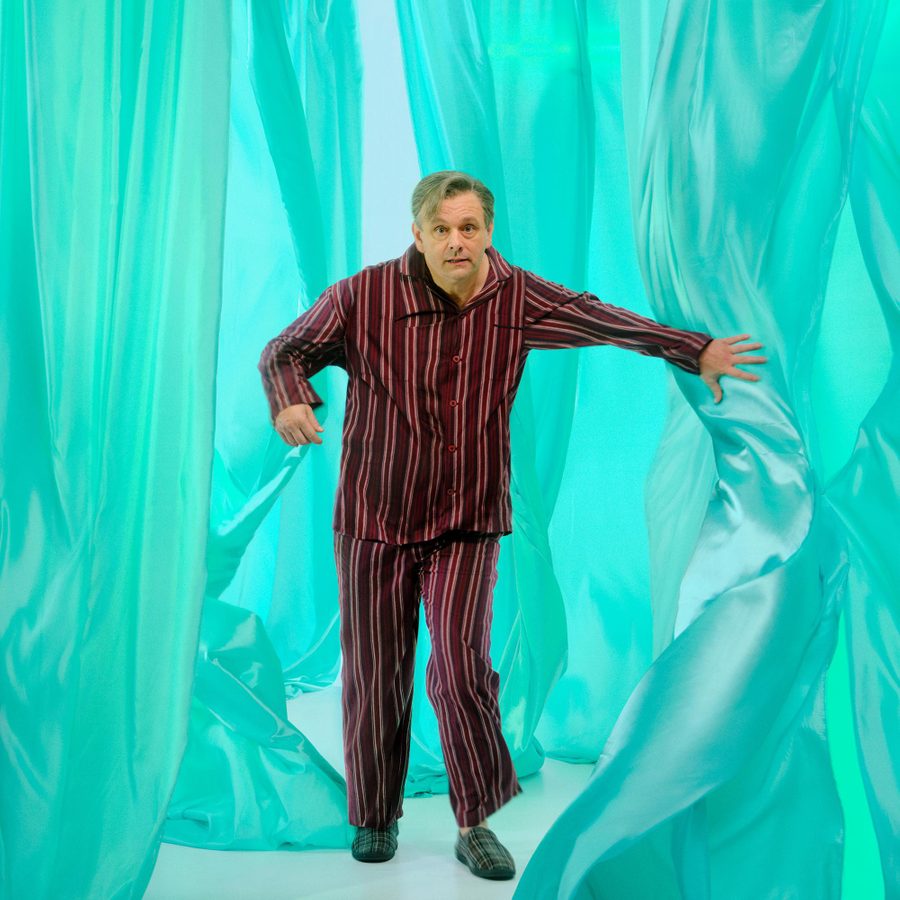 A man wearing red and black striped pajamas and plaid slippers is moving through flowing, translucent teal curtains. He extends his left arm forward, to part one of the curtains. The background consists of soft, undulating teal curtain layers, creating an abstract, dreamy environment.