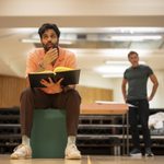 A man in an orange shirt and brown pants holds a script and sits on a green block, looking thoughtful. Another man in a green shirt and jeans stands in the background with his hands on his hips. They appear to be in a brightly lit rehearsal space with wooden walls.