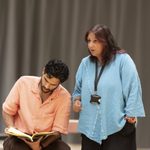 A person with a beard in a peach shirt is sitting and looking at a book, while another person with long hair in a blue blouse stands beside them, speaking. Both are in an indoor setting with a neutral background.