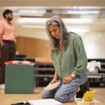 A woman with gray hair, wearing a green shirt and jeans, kneels on the floor with a notepad and pen, intently writing. She has glasses resting on her head. In the background, a man in a pink shirt and brown pants stands, slightly blurred against a wooden wall.