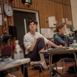 A person with short hair, wearing a white t-shirt, sat with their feet up on a chair in a rehearsal room. They are sat at a table and looking to their right. Various books, water bottles, and other items are on the table in front of them.