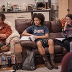 Three people sit on couches in a rehearsal room, engaged in conversation. One person on the left is holding a script, the middle person is wearing a blue graphic t-shirt, and the person on the right is laughing. Various water bottles and bags are on the floor by their feet.