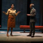 Two people on a stage. The woman on the left is gesturing with both hands and wears a yellow blouse and brown skirt, while the man on the right, dressed in a gray suit, looks at her. Two benches are in the background.