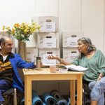 Two people sit and converse at a wooden table. Behind them are stacked boxes labeled with various names. Yellow flowers in a vase and a few rolled-up yoga mats are also visible. The setting appears casual and friendly, possibly a relaxed meeting or workshop setup.