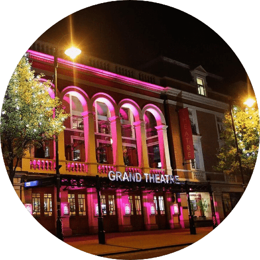 A night view of the Grand Theatre illuminated with pink lights. The historic building features large, arched windows and ornate architectural details. Street lamps and trees frame the scene.