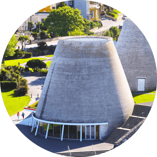 Circular building with a unique, conical structure and large entrance windows, set in a lush green area with trees and pathways. The image captures the building from an elevated perspective, illuminating the detailed texture of the structure in natural light.