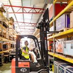 A man operates a black and red electric forklift in a well-organized warehouse with high shelves stocked with various cases and containers. He wears a high-visibility vest and sits in the forklift lifting equipment to the shelves. The warehouse has a metal roof and supports.