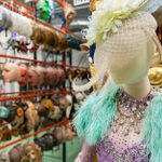 A mannequin in a glamorous purple dress with sparkling beads, a green fur stole, and a fancy white hat with feathers stands in the foreground. Behind it, shelves are filled with various masks and hats, creating a colorful and theatrical atmosphere.