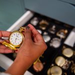 A pair of hands holding an open gold pocket watch. In the background, a drawer filled with various watches and pocket watches in different colors and styles is visible. The drawer is part of a light-colored piece of furniture.