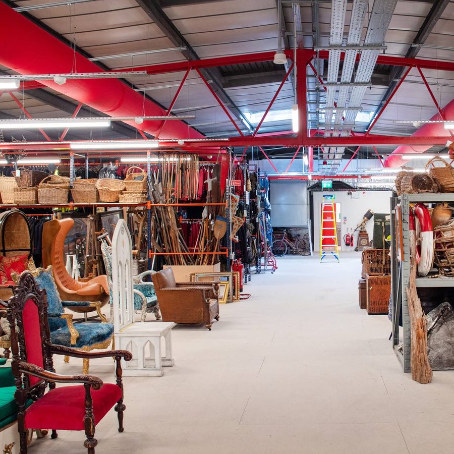 A warehouse filled with assorted vintage furniture, baskets, and various items. Red overhead pipes are visible along with a ladder in the background. Shelves are stocked with a variety of eclectic objects including chairs, statues, and decorative items.