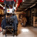 A storage room filled with various military garments and accessories, including uniforms, belts, and gas masks hanging on racks. Shelves in the background hold more equipment and clothing. A ladder is positioned nearby, and the room is brightly lit by fluorescent lights.