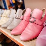 A row of vintage shoes in pastel colors, including shades of pink, white, and beige, displayed on a shelf in a store. Each pair features a shiny, ornate buckle detail on the top. The background shows more shelves and a variety of other shoes.