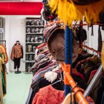 A storage room filled with theatrical costumes is shown. Rows of colorful, period dresses hang on racks on both sides of a narrow, green-tiled aisle. At the end of the aisle is a mannequin dressed in an outfit, and shelves with numerous helmets line the back wall.