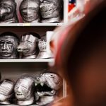 A collection of medieval helmets and masks displayed on shelves. The headgear features various designs, including human faces, animals, and abstract shapes. The foreground is slightly blurred, highlighting the intricate details of the items on the shelves.