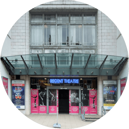 Image of the entrance to Regent Theatre with glass canopy and neon sign. Posters for upcoming shows and events are displayed on either side of the entrance. The building has a tiled façade and large windows above the entrance.