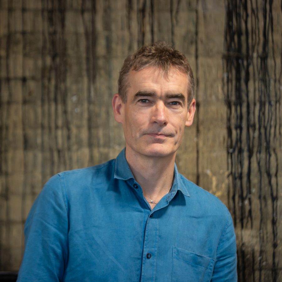 A man with short brown hair wearing a blue button-up shirt stands in front of a textured, abstract background with vertical lines. He has a neutral facial expression and is looking directly at the camera.