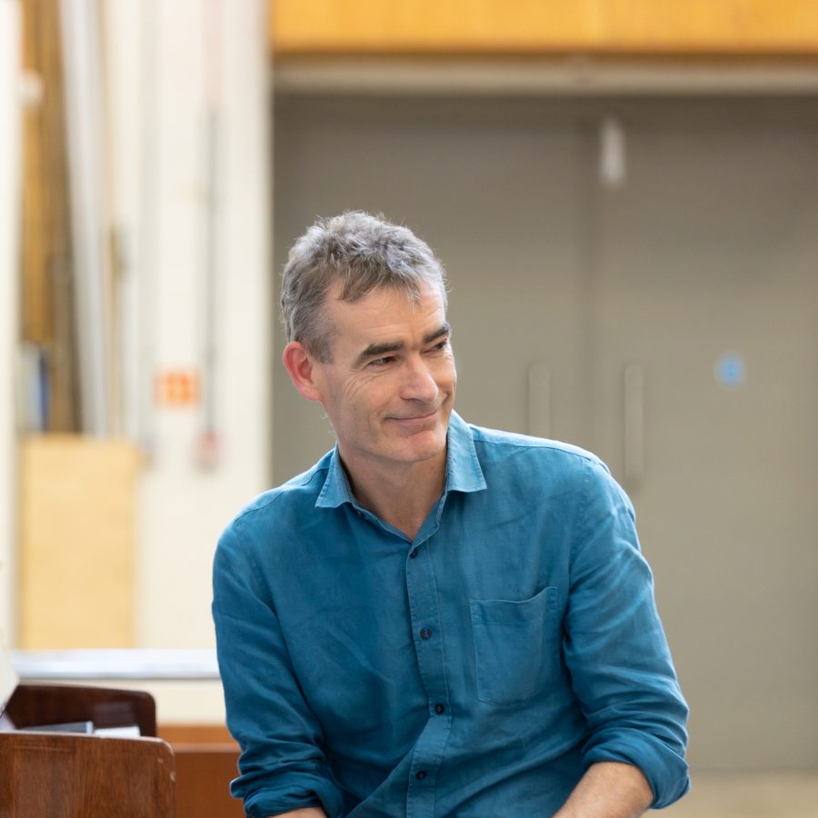 A man with gray hair, wearing a blue button-up shirt, is smiling while sitting indoors. The background is slightly blurred and appears to be a room with beige walls and a closed door.