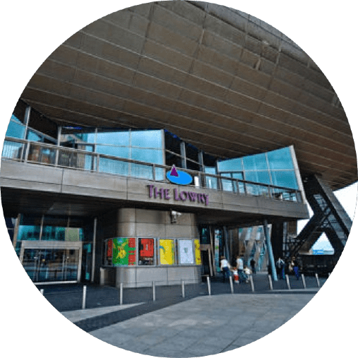 The image depicts the exterior of The Lowry, a contemporary glass and steel building with an overhanging structure. The entrance has large glass windows and a balcony above it. Several people are walking near the entrance and colorful posters are displayed.