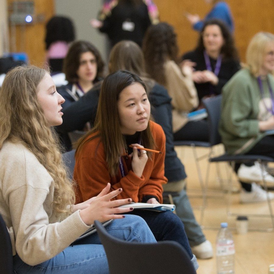 A group of diverse people sitting in a room, engaged in conversation. Three women in the foreground are sitting and talking, holding notebooks and pens. Others in the background are talking and writing, suggesting an interactive workshop setting.