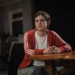 A young woman with a thoughtful expression sits at a wooden table, wearing a red jacket with white stripes over a white shirt. The background is dimly lit with blurred details.