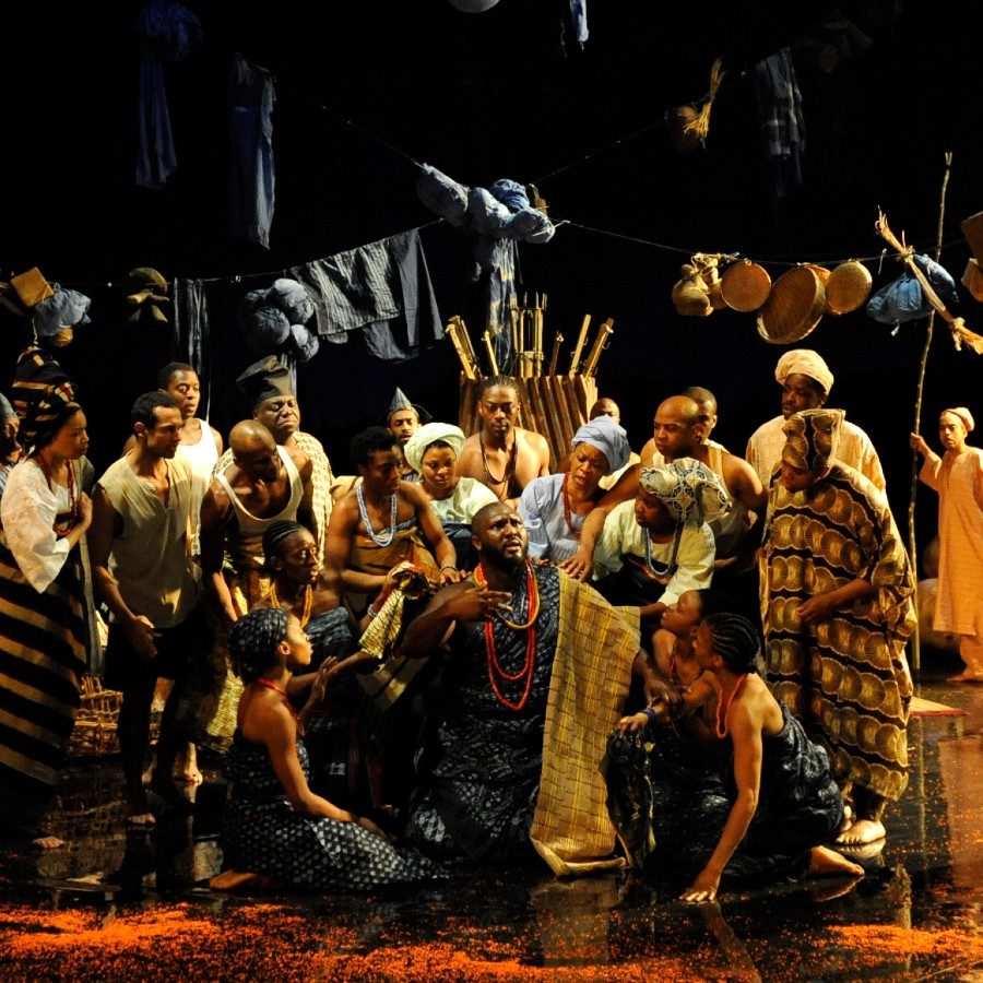 A group of people on stage in colourful attire, set against a dark background. They are surrounded by props like baskets and cloths hanging above them. The scene conveys a sense of community or celebration.