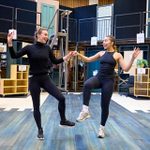 Two women in athletic wear practice a dance routine on a blue wooden floor, smiling and holding hands. In the background, a woman in a pink skirt watches, with various stage props and doors around them in a rehearsal space.