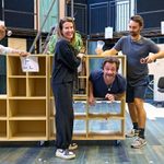 Four people having fun with an empty wooden shelf on wheels. A man is leaning through one of the shelf openings while another man points a torch at him. Another man holds a torch and an empty cup over the man leaning through the shelf. A woman is stood to the side laughing. Behind them is a bare bones theatre set with two levels, and multiple doors.