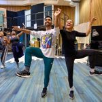 A group of performers rehearse a dance in a studio with wooden walls and a piano. They are smiling and wearing casual dance attire, striking a high-energy pose with one arm up and one knee lifted.