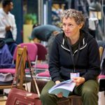 A person with short curly hair sits on a chair holding a script, wearing glasses and a hoodie. The background includes chairs, a hat, and people engaged in different activities.