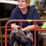 A person with short hair leans on a red railing, looking outward with a thoughtful expression. In front of them is a dinosaur skull model.