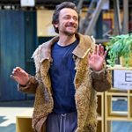 A man in casual clothing and a rugged, furry coat smiles and gestures with his hand in a rehearsal space. Behind him is a bare bones theatre set with two levels, and some wooden shelves.