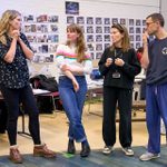 Four people stand in a discussion in a room filled with photos and documents of costume and set designs on the walls. The woman on the left is speaking, and the other three people are listening to her intently.