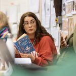 A woman with glasses and a red sweater holds a book titled 