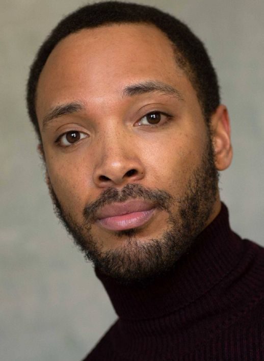 A person with short black hair, a closely trimmed beard, and mustache gazes slightly off-camera. They are wearing a dark-colored turtleneck sweater against a plain, soft-focused background.