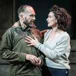 A man (Ralph Fiennes) with a beard and short hair, wearing a khaki jacket with camo sleeves, stands on stage with a woman (Indira Varma) with short, curly hair in a grey cardigan. They are stood close together, her hand on his chest, staring at each other with serious expressions. The background is dimly lit with concrete steps visible.