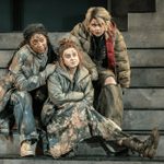 Three women with dishevelled hair and ripped clothes sit closely together on concrete steps on a stage. They are all looking in the same direction with raised eyebrows. The stage is brightly lit.