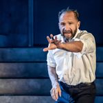 A man (Ralph Fiennes) with a beard and short hair, wearing a white shirt with rolled-up sleeves, stands on stage. He extends one arm forward, with a serious expression. The background is dimly lit with concrete steps visible.