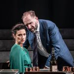 A man (Ralph Fiennes) with a beard and short hair, wearing a blue jacket and a tie, stands on stage with a woman (Indira Varma) with short, curly hair in a blue dress. She is sat and he is stood close to her, leaning over a table, staring at empty space with serious expressions. The background is dimly lit with concrete steps visible.