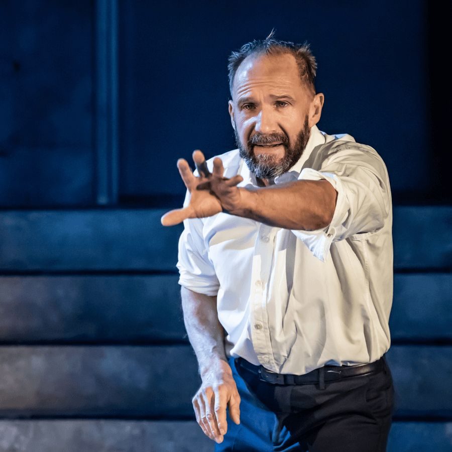 A man (Ralph Fiennes) with a beard and short hair, wearing a white shirt with rolled-up sleeves, stands on stage. He extends one arm forward, with a serious expression. The background is dimly lit with concrete steps visible.